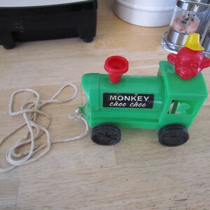 May include: A green toy train with a monkey face on the front. The train has black wheels and a red whistle. The train is attached to a white string.