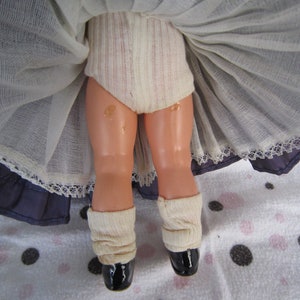 Pu&ograve; includere: Una bambola vintage con un bordo di pizzo bianco su una gonna blu, che indossa calze bianche e scarpe nere.