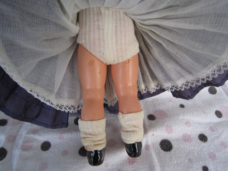 Pu&ograve; includere: Una bambola vintage con un bordo di pizzo bianco su una gonna blu, che indossa calze bianche e scarpe nere.