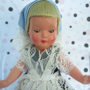 Pu&ograve; includere: Una bambola di porcellana vintage con capelli biondi, occhi azzurri e un vestito di pizzo bianco. La bambola indossa un berretto blu e ha un incarnato rosato.