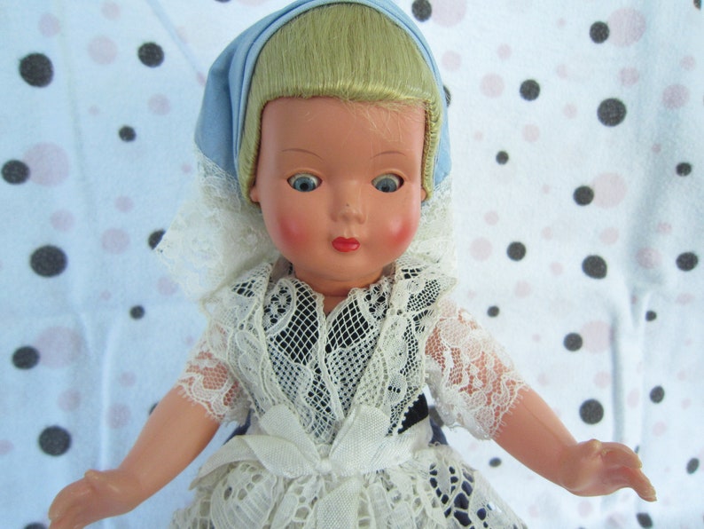Pu&ograve; includere: Una bambola di porcellana vintage con capelli biondi, occhi azzurri e un vestito di pizzo bianco. La bambola indossa un berretto blu e ha un incarnato rosato.