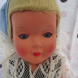 Pu&ograve; includere: Una bambola di porcellana vintage con capelli biondi, occhi azzurri e guance rosate. Indossa un colletto di pizzo bianco e un berretto blu e bianco.