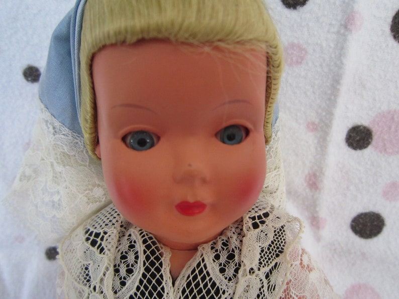 Pu&ograve; includere: Una bambola di porcellana vintage con capelli biondi, occhi azzurri e guance rosate. Indossa un colletto di pizzo bianco e un berretto blu e bianco.