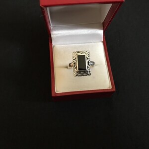 May include: A silver ring with a black rectangular stone set in a decorative frame. The ring is in a red velvet box.