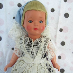 Pu&ograve; includere: Una bambola di porcellana vintage con capelli biondi, vestita con un abito di pizzo bianco e un foulard blu e bianco per la testa. La bambola &egrave; posizionata su uno sfondo bianco con pois neri e rosa.