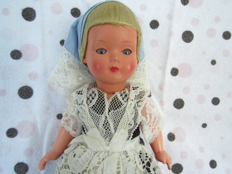 Pu&ograve; includere: Una bambola di porcellana vintage con capelli biondi, vestita con un abito di pizzo bianco e un foulard blu e bianco per la testa. La bambola &egrave; posizionata su uno sfondo bianco con pois neri e rosa.