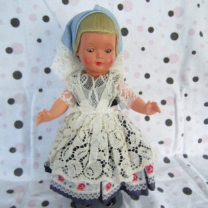 Pu&ograve; includere: Una bambola vintage che indossa un abito di pizzo bianco con dettagli floreali blu e rosa. La bambola ha i capelli biondi e gli occhi azzurri e indossa un foulard blu e bianco.