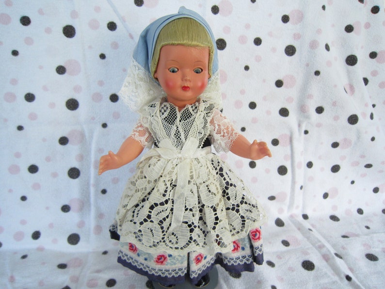 Pu&ograve; includere: Una bambola vintage che indossa un abito di pizzo bianco con dettagli floreali blu e rosa. La bambola ha i capelli biondi e gli occhi azzurri e indossa un foulard blu e bianco.