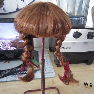 May include: A brown wig with two braids tied with red ribbons. The wig is on a red metal stand. Wig #5.