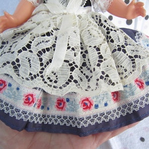 Pu&ograve; includere: Un vestito da bambola con un'applicazione di pizzo bianco, un bordo floreale blu e bianco e una base in tessuto blu.