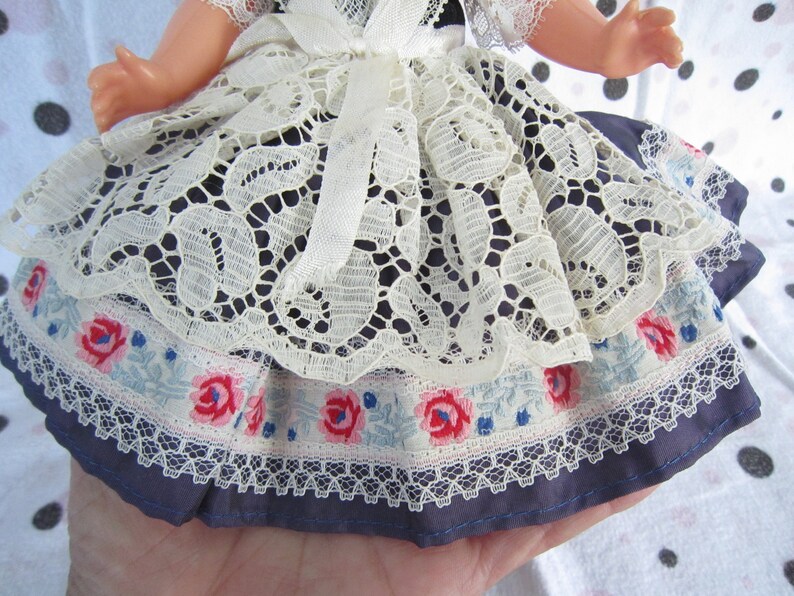 Pu&ograve; includere: Un vestito da bambola con un'applicazione di pizzo bianco, un bordo floreale blu e bianco e una base in tessuto blu.