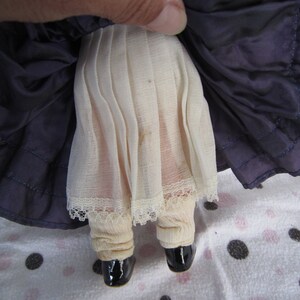 Pu&ograve; includere: Una bambola di porcellana vintage che indossa un vestito blu con un bordo di pizzo bianco e un sottogonna bianco. La bambola indossa scarpe nere.