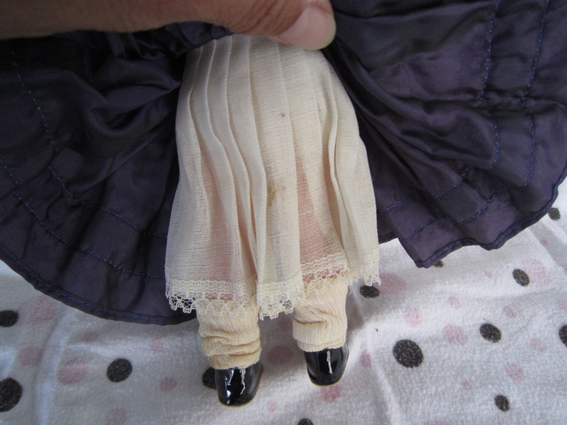 Pu&ograve; includere: Una bambola di porcellana vintage che indossa un vestito blu con un bordo di pizzo bianco e un sottogonna bianco. La bambola indossa scarpe nere.
