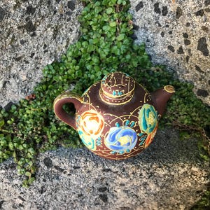 May include: A small, decorative brown teapot with a lid, handle, and spout. The teapot is adorned with colorful floral designs in blue, orange, and green, outlined in gold. It rests on a stone surface with green foliage.