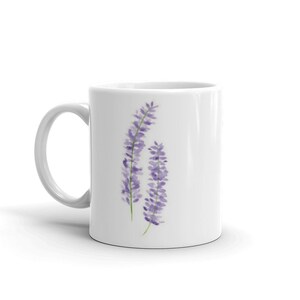May include: White ceramic mug featuring a watercolor illustration of two lavender sprigs. The sprigs are a soft purple with green stems, set against the white background of the mug. The mug has a curved handle.