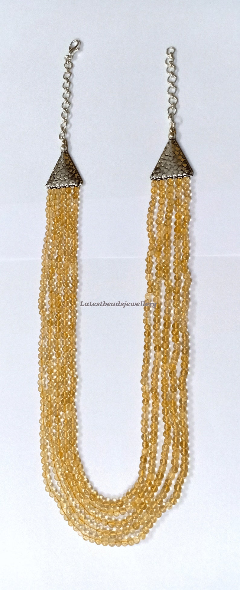 May include: A multi-strand necklace made of light brown beads with silver triangle end caps. The necklace is on a white background.