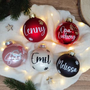 Pu&ograve; includere: Una collezione di cinque ornamenti natalizi in vari colori: rosso, rosa, bianco e nero. Ogni ornamento presenta un nome in corsivo bianco, come "Lenny" e "Melissa", insieme a disegni di fiocchi di neve e impronte di zampe. Gli ornamenti sono su un tessuto bianco.