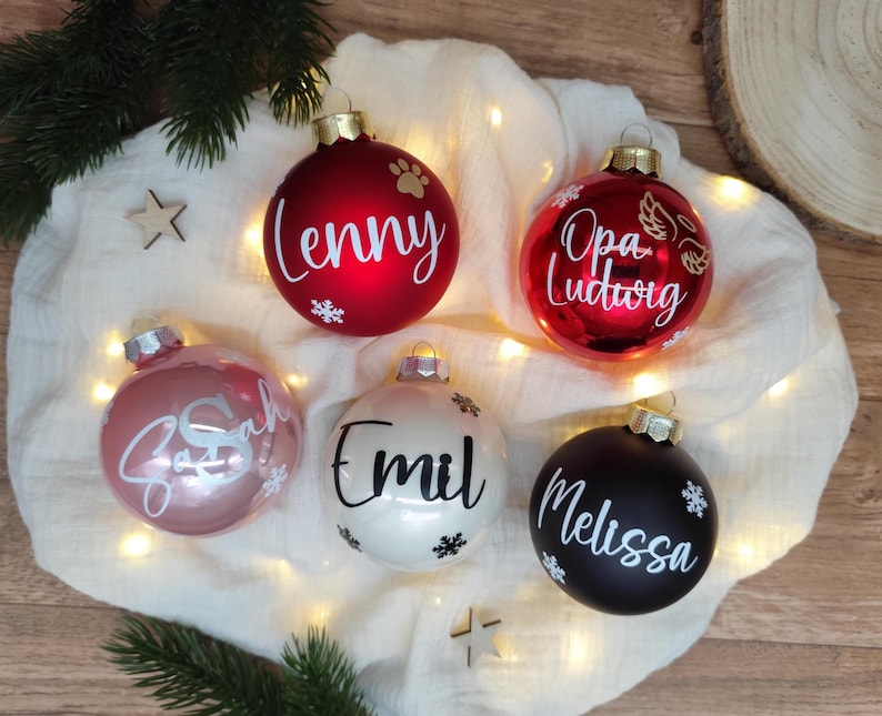 Pu&ograve; includere: Una collezione di cinque ornamenti natalizi in vari colori: rosso, rosa, bianco e nero. Ogni ornamento presenta un nome in corsivo bianco, come "Lenny" e "Melissa", insieme a disegni di fiocchi di neve e impronte di zampe. Gli ornamenti sono su un tessuto bianco.