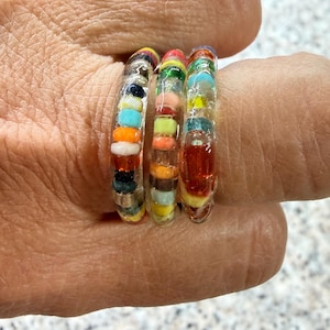 May include: Three colorful beaded rings. Each ring is clear with a variety of small, colorful beads in shades of red, orange, yellow, green, blue, and black. The rings are worn on a finger.