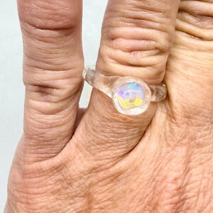May include: A clear, iridescent glass ring with a round, colorful center stone. The ring is worn on a finger, showcasing its delicate design and the play of light within the stone. The ring's band is also clear, blending with the skin.