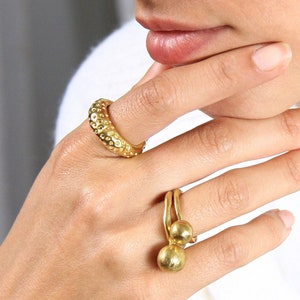 Peut inclure: Une bague en or avec deux boules rondes attachées à la bande. La bague est portée sur le majeur d'une main. La main porte également une bague en or avec une surface texturée sur l'index.