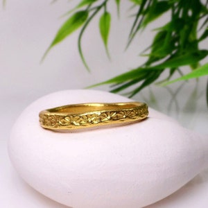 May include: A thin gold band ring with a floral design. The ring is on a white surface.