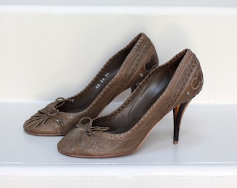 May include: A pair of brown leather heeled shoes with a decorative stitch pattern and a bow detail on the toe.