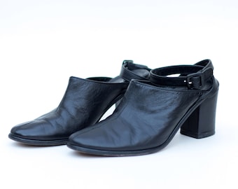 Puede incluir: Un par de zapatos de tacón de cuero negro con correas de tobillo. Los zapatos tienen una punta puntiaguda y un tacón grueso.