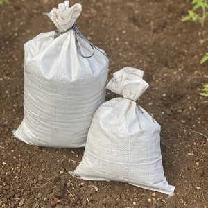 May include: Two white burlap sacks, one larger than the other, are sitting on a brown dirt surface. The sacks are tied with a black string.
