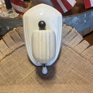 May include: A wall-mounted cream-colored ceramic kitchen tool featuring a vertical shield-shaped backplate with stepped edges, a central fluted container, a small black knob at the top, and a white handle extending from the bottom center.