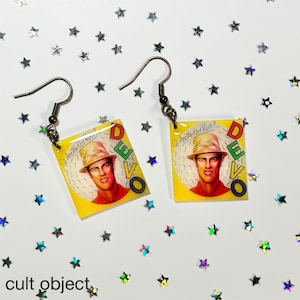 May include: A pair of dangle earrings featuring a square-shaped charm with a colorful image of a man wearing a hat and the text "Are We Not Men? DEVO".
