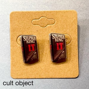 May include: A pair of earrings featuring the cover of Stephen King's novel "IT" on a brown card with the text "cult object".