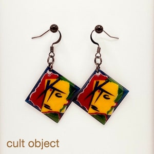May include: A pair of abstract art earrings. Each earring features a yellow face with red hair and a blue border. The earrings are diamond-shaped and hang from black hooks. The text "cult object" is at the bottom.
