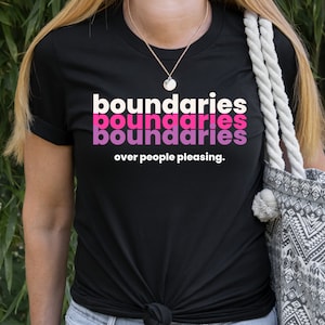 May include: A black t-shirt with the words "boundaries boundaries boundaries" in pink and white text. The text "over people pleasing." is printed below the words "boundaries".