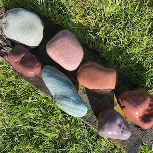 May include: A collection of seven smooth, polished river rocks in various shades of brown, gray, and green. The rocks are arranged in a line on a piece of weathered wood.
