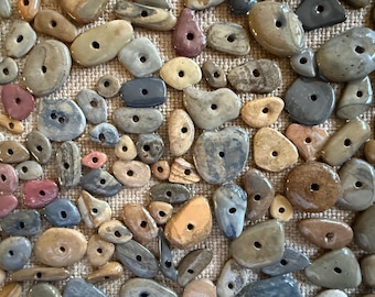 Puede incluir: Una colección de piedras lisas perforadas en varias formas y colores, incluyendo beige, gris, rosa y marrón. Cada piedra tiene un solo agujero, lo que sugiere que son cuentas o elementos decorativos. Las piedras están dispuestas sobre una superficie texturizada de color claro.