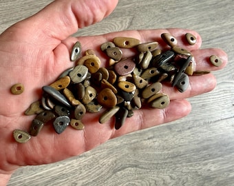 Puede incluir: Una mano sostiene una colección de pequeñas cuentas de piedra natural. Las cuentas varían en forma, incluyendo formas redondeadas, ovaladas y alargadas, con un agujero central para ensartar. Los colores van del beige y marrón al gris y negro, con algunos patrones rayados.