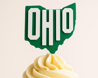 May include: A cupcake with a swirl of yellow frosting, topped with a green and white Ohio state-shaped decoration that spells out "OHIO". The cupcake sits in a white paper liner against a light background.