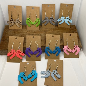 May include: A display of 10 pairs of dinosaur skull earrings. The earrings are made of acrylic and come in a variety of colors, including pink, blue, green, purple, and gray.