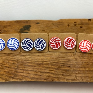 May include: Six pairs of volleyball-themed earrings on brown card stock. The earrings are white with blue or red volleyball designs.