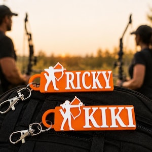May include: Two orange rectangular keychains with white lettering and a white silhouette of an archer. One keychain features the name RICKY and the other features the name KIKI. Each keychain includes a silver metal lobster clasp attachment.