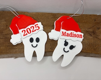 May include: Two white tooth-shaped Christmas ornaments with red Santa hats. One ornament has the year "2025" and the other has the name "Madison" in red. Both have black eyes and a smiling mouth. Each ornament has a silver string for hanging.