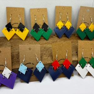 May include: Assortment of handmade earrings featuring two layered geometric shapes in various colors, including yellow, green, blue, red, purple, white, and black. Each pair is displayed on a brown card with the text "Handmade by" and a name.