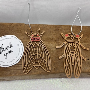 May include: Two wooden cicada ornaments with red accents. One ornament has the year 2023 written on it. The ornaments are hanging from a string.