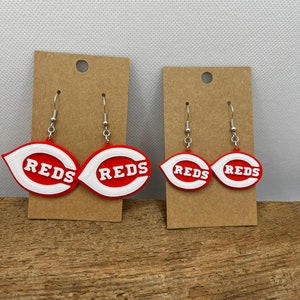 May include: A pair of red and white Cincinnati Reds earrings. The earrings are shaped like the team's logo, a red 'C' with a white outline. The earrings are hanging from silver hooks.