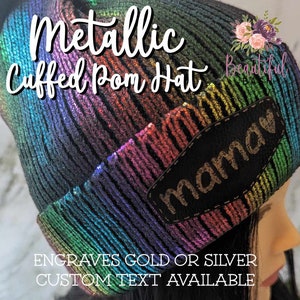 May include: Metallic rainbow knit beanie with a black pom pom and a black patch that says "mama" in gold lettering. The beanie is cuffed and has a metallic sheen. The text "Engraves Gold or Silver Custom Text Available" is written below the beanie.