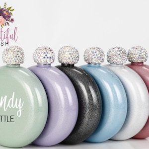May include: Six round, metallic hip flasks with glitter finishes in various colors, including green, purple, black, blue, white, and pink. Each flask has a clear, crystal-like stopper with rhinestones. The green flask has the text "wendy LITTLE" printed on it.