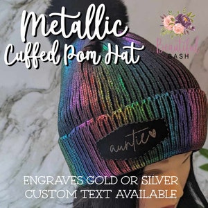May include: Metallic rainbow iridescent cuffed beanie with a faux fur pom pom and a black patch that says "auntie" with a heart. The beanie is available with engraved gold or silver text.