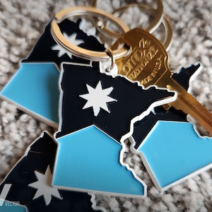 May include: A keychain featuring three blue and black outline shapes of the state of Minnesota with a white star in the center of each shape. A gold key with the text "Otto Z.  Nationalize Made in U.S.A." is attached to the keychain.