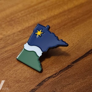 May include: A pin in the shape of the state of Minnesota, with a navy blue top section featuring a yellow star, a white middle section, and a green bottom section. The pin is on a wooden surface.